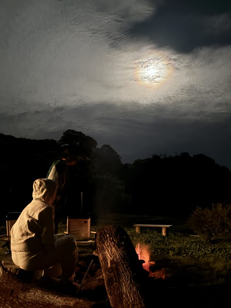 夜空に浮かぶ満月と焚き火を囲む人たちの後ろ姿