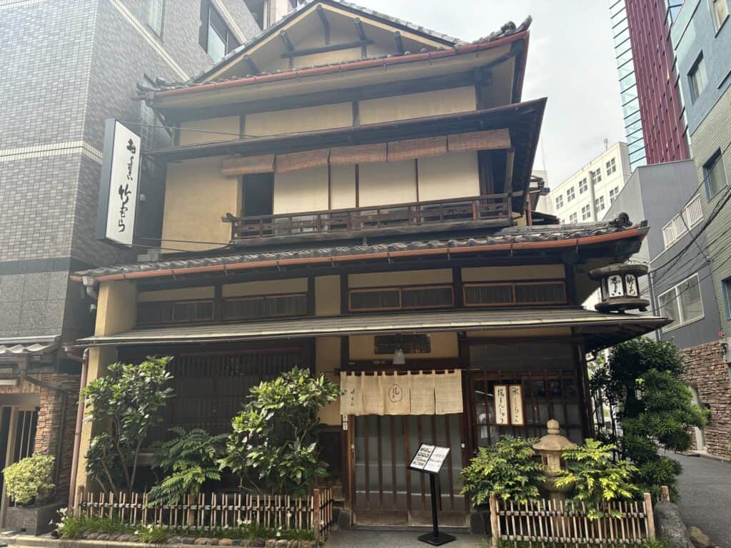 「竹むら」の白い縦看板が立つ、木造3階建ての外観。入口にのれん、右手に吊り提灯と石灯籠、手前に植え込みと立て看板が見える