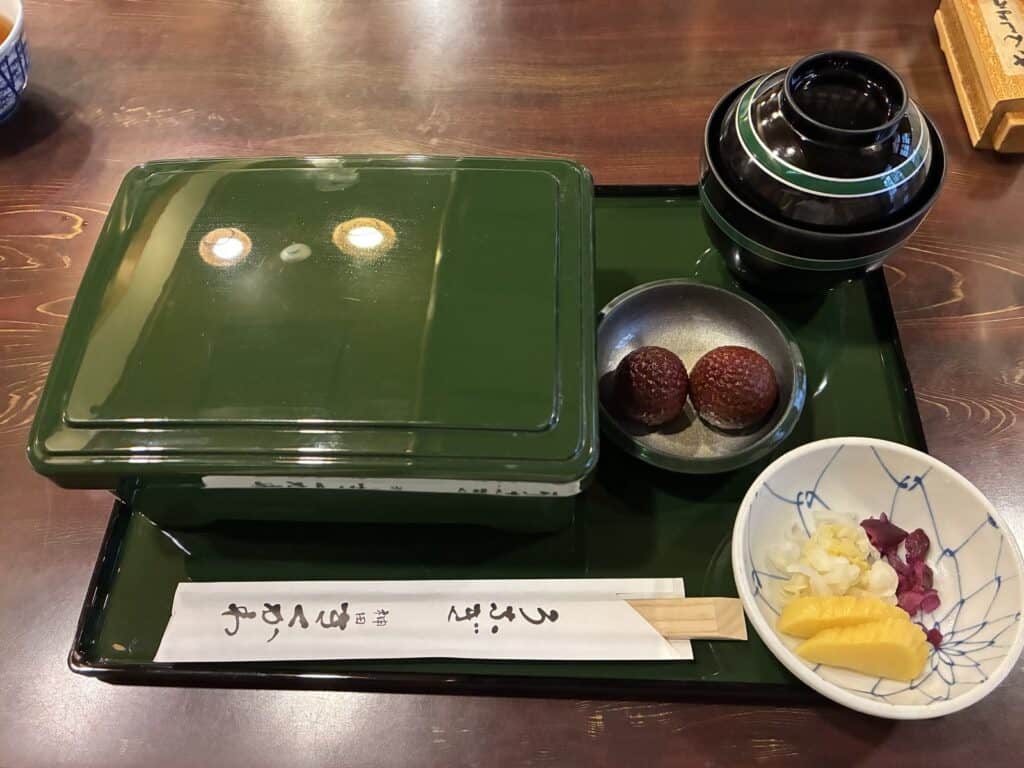 緑のお盆にのった、フタの閉まったお重と吸い物、香の物、ガラスの器に入ったライチ2粒