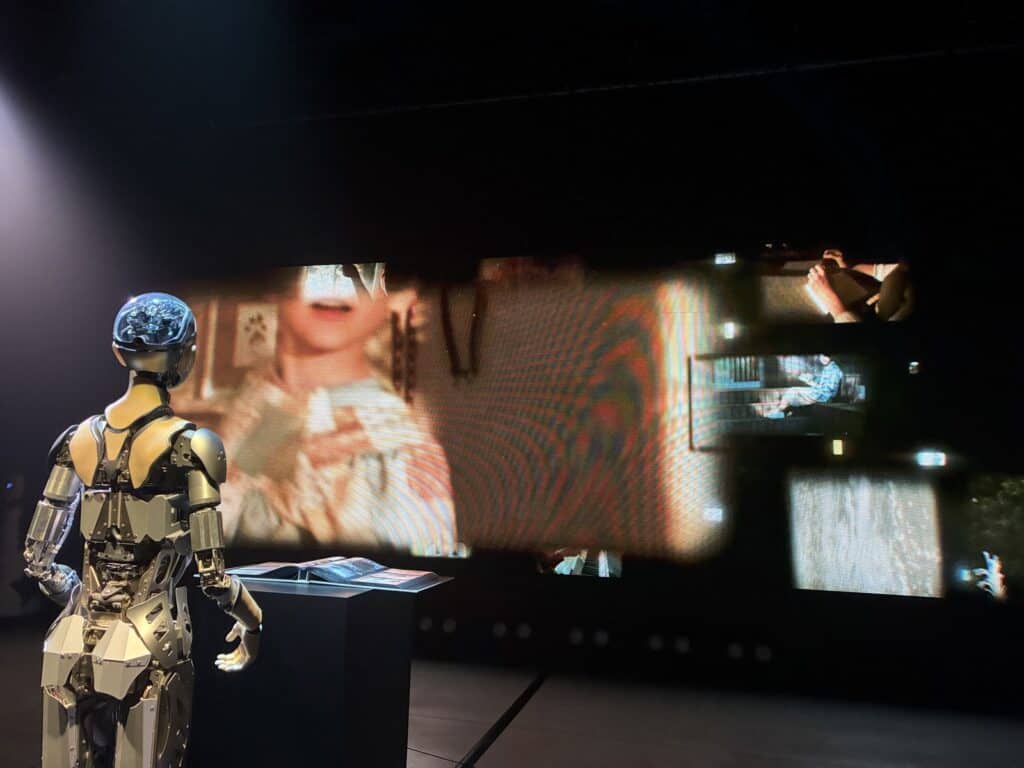 暗い展示室に立つアンドロイドと、壁に映し出された映像