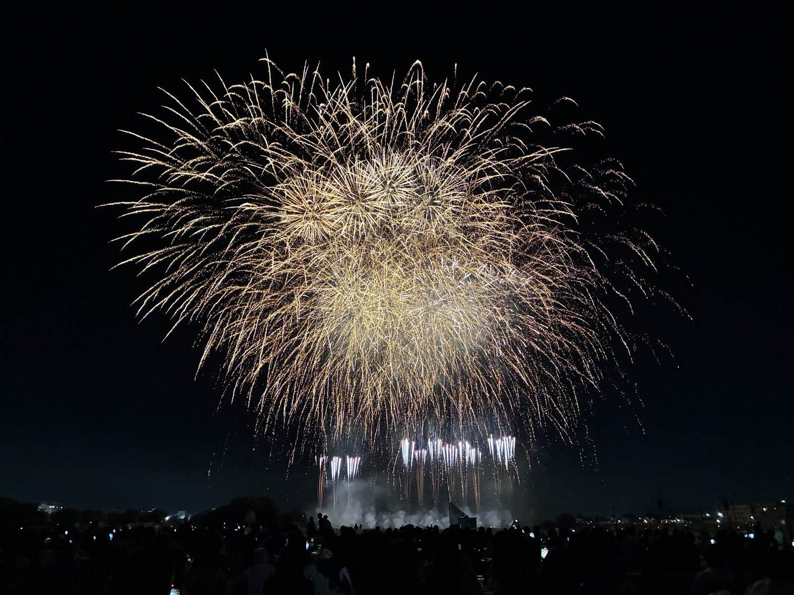 金色の大きな花火が夜空に開き、下で白い光が噴き上がっている